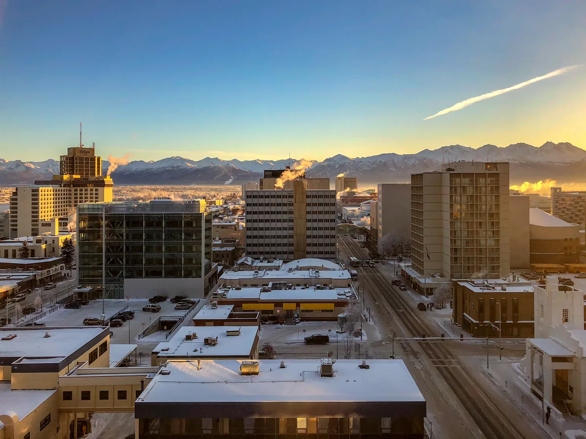 The sun sets on Downtown Anchorage on a cold, quiet day in February, with the Chugach Mountain Range just below the horizon above snow-cover — Anchorage, AK